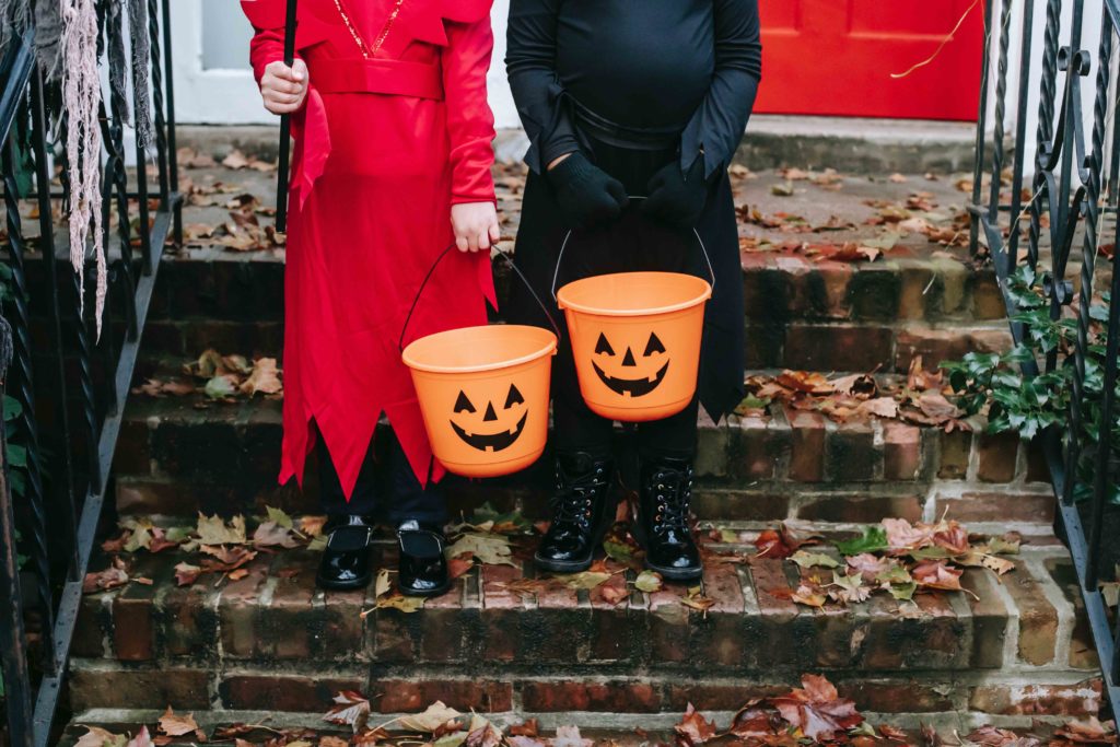two kids in costume standing on a brick front porch with baskets shaped like pumpkins