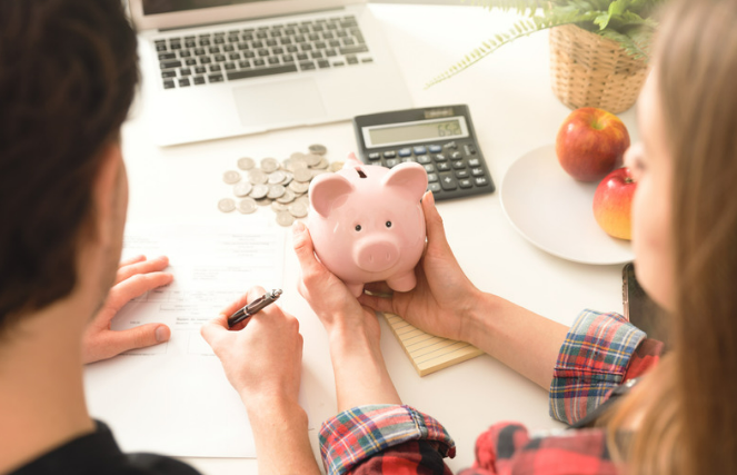 A couple does some budget and financing with an apple beside and a calculator in the table. The girl was holding their piggy bank while the man holding a pen did some financing as a first-time homebuyer
